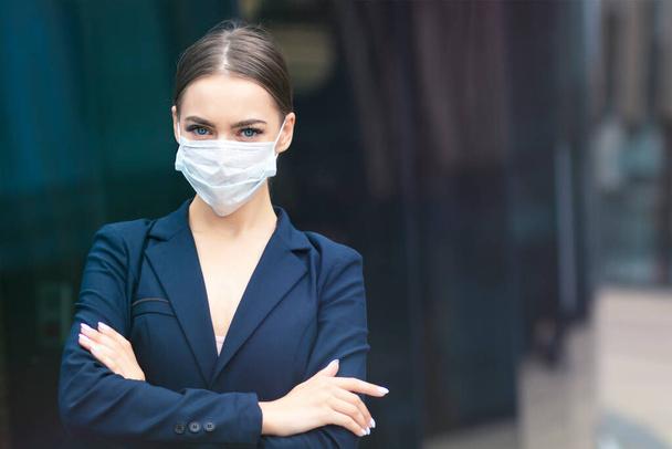 Beautiful serious business woman standing outdoors with her hands crossed, young gorgeous pretty girl in medical sterile protective mask against coronavirus, epidemic covid-19. Healthcare at job, work - Foto, Imagen