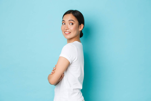 Kokettierendes hübsches asiatisches Mädchen in weißem T-Shirt, das im Profil steht und mit neugierig lächelndem Gesicht nach rechts abbiegt, sich etwas Interessantes ansieht, gutes Promo-Angebot findet, vor blauem Hintergrund steht - Foto, Bild