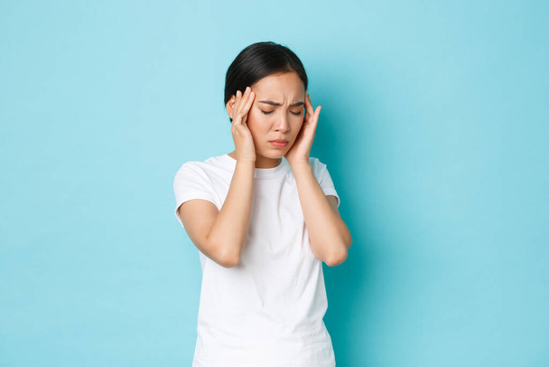 Side view of bothered tired asian girl grimacing, touching head and complaining on headache, having pain in head, migraine of feeling dizzy, standing blue background sick - Photo, Image