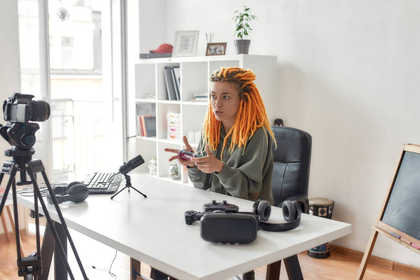Female technology blogger with dreadlocks holding game controller joystick while recording video review of new gadgets using microphone at home - Zdjęcie, obraz