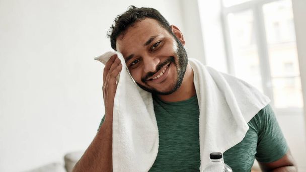 Sweaty from exercising. Portrait of young man smiling at camera, wiping sweat with towel while resting after training workout at home. Sport, healthy lifestyle - Фото, зображення