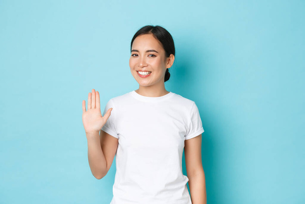 Smiling friendly-looking asian girl in white t-shirt waving hand in hello gesture, saying hi, greeting person and standing blue background happy, glad to meet someone - Photo, Image