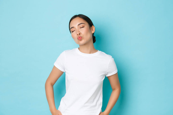 Silly and cute asian girl close eyes and dreamy pouting, waiting or imaging romantic kiss, looking lovely over blue background in casual white t-shirt, lifestyle and people concept - Photo, Image