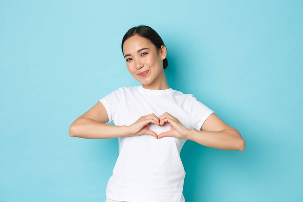 Lovely asian girl in white t-shirt showing sympathy, express care and tender feelings towards person, showing heart gesture and smiling silly at camera, standing blue background - Фото, изображение