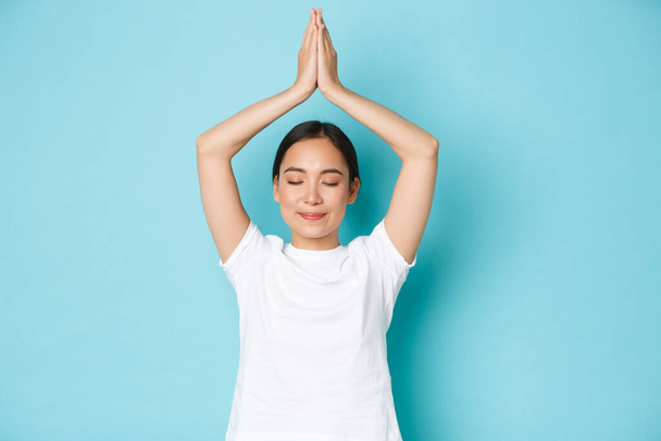 Portret van gelukkig en vredig, ontspannen lachend Aziatisch meisje dat zorgt voor haar geestelijke gezondheid, yoga of meditatie beoefent, ogen sluit en handen omhoog houdt in asana pose, blauwe achtergrond - Foto, afbeelding