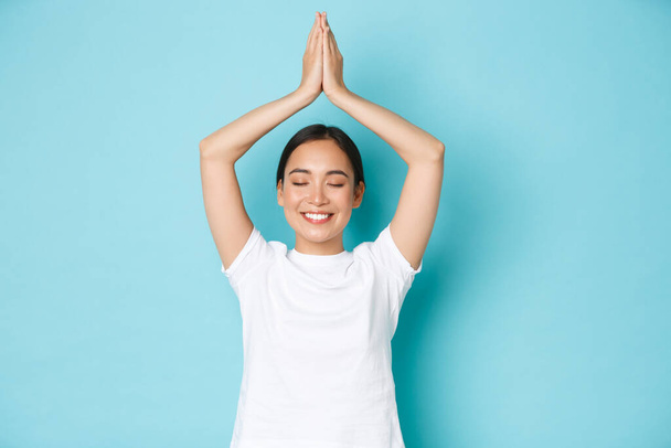 Portré boldog és békés, nyugodt mosolygós ázsiai lány vigyáz a mentális egészség, gyakorlása jóga vagy meditáció, csukott szemmel, és emelje fel a kezét a feje fölé ázsiai pózban, kék háttér - Fotó, kép
