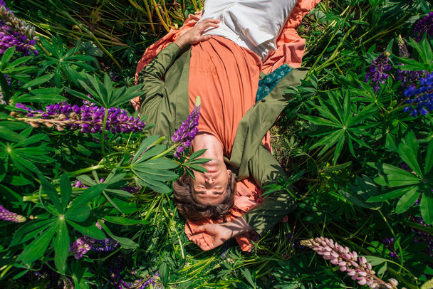 Tall handsome man in a green jacket lying among the lupines flowers. Top view. - 写真・画像