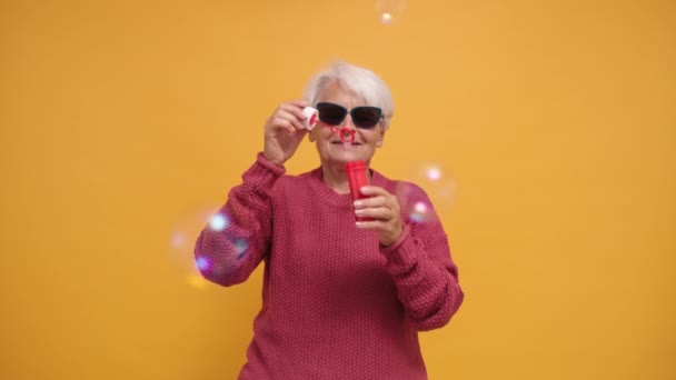 Μοντέρνα γιαγιά με γυαλιά ηλίου και μακρυμάνικο πουλόβερ που φυσάει σαπουνόφουσκες. Μεμονωμένα - Πλάνα, βίντεο