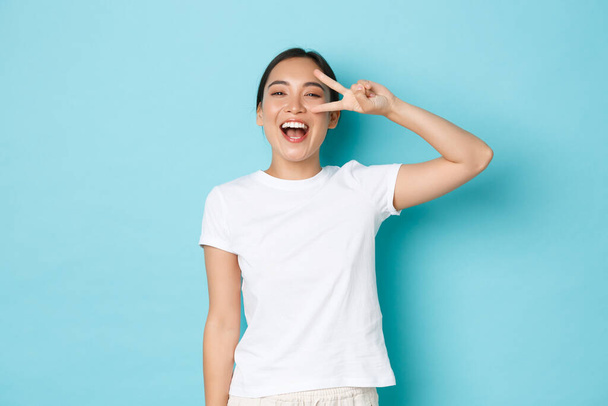 Lifestyle, την ομορφιά και τις γυναίκες έννοια. Χαρούμενη όμορφη Ασιάτισσα με λευκό t-shirt που κοιτάζει αισιόδοξα και Kawaii, που δείχνει την ειρηνική χειρονομία πάνω από το μάτι και χαμογελά ευρέως, στέκεται μπλε φόντο - Φωτογραφία, εικόνα