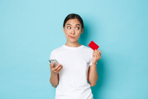 Sähköinen kaupankäynti, ostokset ja elämäntapa. Epämääräinen huomaavainen aasialainen tyttö valkoisessa t-paidassa, katselee pois ajattelemalla pitäen luottokorttia ja älypuhelinta, tehden online-tilauksen - Valokuva, kuva