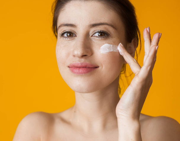Caucasian woman with freckles applying anti aging cream on face posing with naked shoulders on yellow studio wall - Foto, imagen