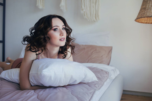 Μια νεαρή γυναίκα με μέικ-απ και μπούκλες στο κρεβάτι στην κρεβατοκάμαρα και αγκαλιάζει το μαξιλάρι χαμογελά - Φωτογραφία, εικόνα