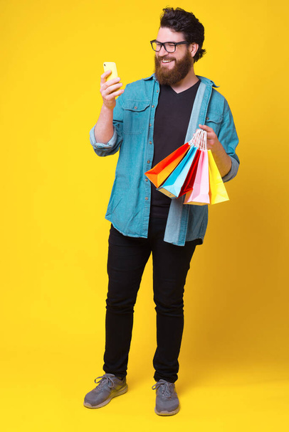 Photo of cheerful hipster man holding shopping bags and using phone - Photo, Image