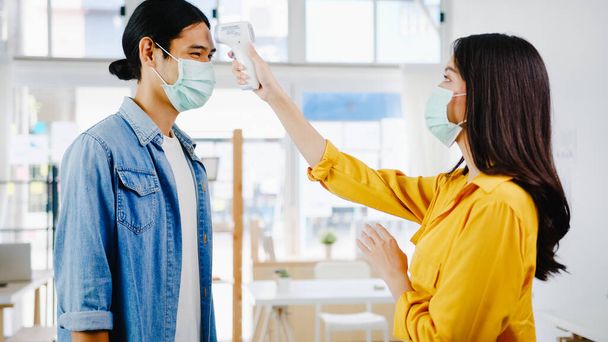 Ázsia női recepciós vezető viselni védő arc maszk használata infravörös hőmérő ellenőrző vagy hőmérséklet pisztoly az ügyfél homlokán, mielőtt hivatalba. Életmód új normális után koronavírus. - Fotó, kép