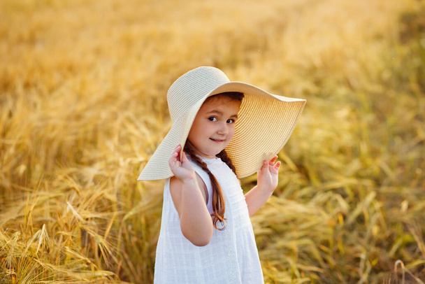 Portret van een mooi meisje in een witte zonnejurk en strohoed op een Gouden tarweveld bij zonsondergang. Meisje geniet van de herfst - Foto, afbeelding