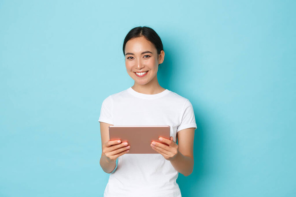 Retrato de sonriente hermosa asiática estudiante trabajando en proyecto, chica usando tableta digital y mirando a la cámara complacido, de pie fondo azul claro, compras en línea - Foto, imagen