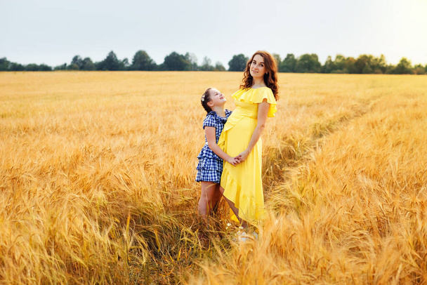 Happy family: a young beautiful pregnant woman with her little cute daughter walking in the wheat orange field on a sunny summer day. Parents and kids relationship. Nature in the country. - Photo, Image