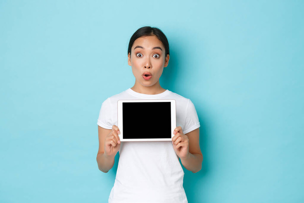 Portret podekscytowanej i zdumionej azjatyckiej dziewczyny dyszącej zdumiony i zdumiony, demonstrujący niesamowity sklep internetowy, pokazujący cyfrowy ekran tabletu i patrzący w kamerę, stojące jasnoniebieskie tło - Zdjęcie, obraz