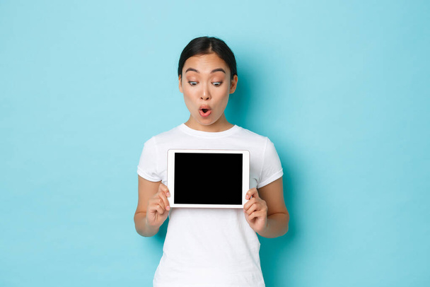 Excited and surprised beautiful asian female in white t-shirt looking at digital tablet screen with astonished, thrilled look, showing chart or demonstrate online shopping site, light blue background - Foto, afbeelding