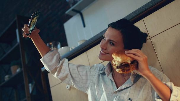 Chica hambrienta comiendo hamburguesa en el suelo. Mujer haciendo foto selfie con hamburguesa. - Foto, Imagen