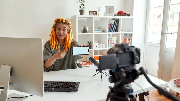 Female technology blogger with dreadlocks looking at camera, showing smartphone with blank screen while recording video blog or vlog in the daytime - Photo, image