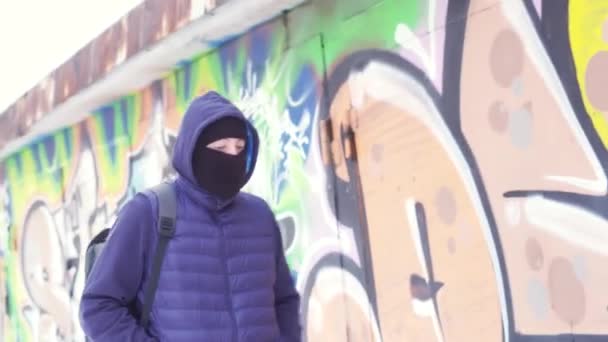 Komoly fiatalember alkalmi ruhában és símaszkban sétál a grafitti falán. Felvétel. Férfi hátizsákkal elrejti az arcát és körülnéz, a vandalizmus fogalma.. - Felvétel, videó