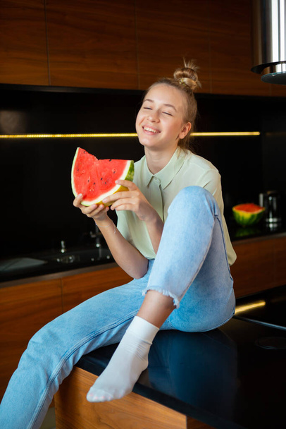 Cute smiling girl sitting with closed eyes with pleasure and holding watermelon in cozy kitchen - Foto, Imagen