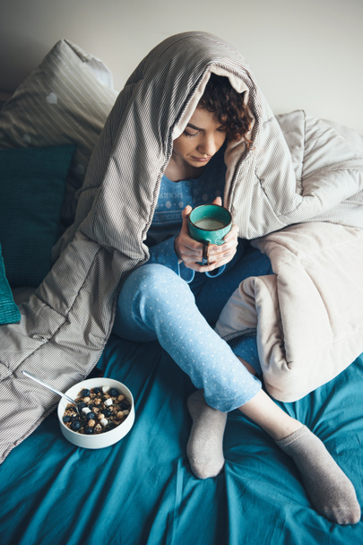Felső nézet fotó egy göndör hajú nő iszik egy csésze tejet, miközben eszik gabonát az ágyban takaróval borított - Fotó, kép