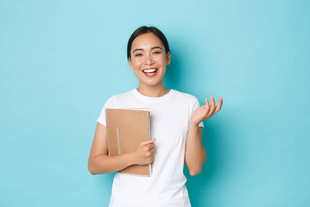 Lifestyle, education and people concept. Cheerful smiling asian female student in white t-shirt, holding notebooks and lively chatting with classmates, gesturing over light blue background - Foto, afbeelding