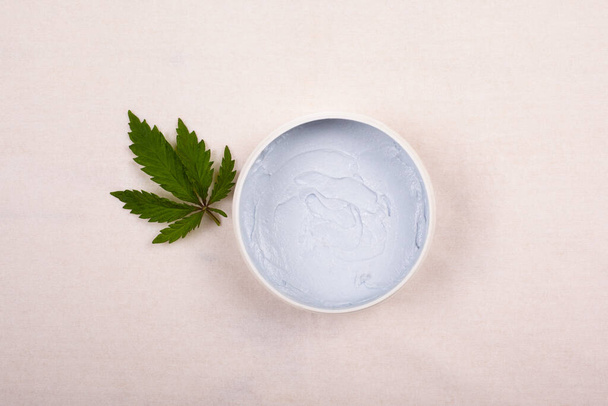 white round jar with a body scrub with a leaf of marijuana on a white background top view, cosmetics, skin care, beauty - Photo, Image