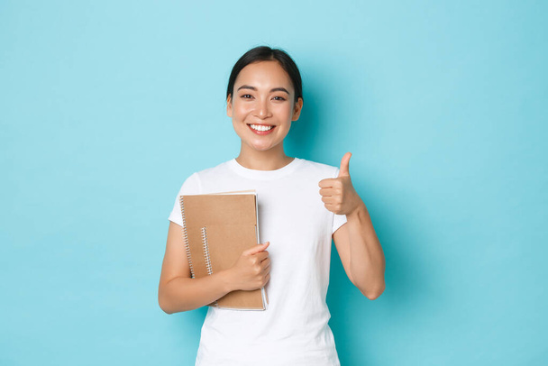 Lifestyle, education and people concept. Portrait of satisfied and happy asian female student in white t-shirt, korean girl showing thumbs-up, carry notebooks, recommend college - Foto, Imagen