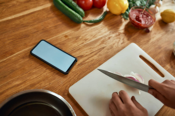 Close up of phone with blank screen on the table. Man cutting onion on chopping board, using smartphone app while cooking dinner. Healthy nutrition, technology concept - Фото, изображение