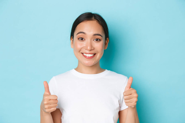 Detailní záběr spokojené a veselé usměvavé asijské dívky v bílém tričku, ukazující palce-up podporující a vypadající spokojeně na fotoaparát, doporučit produkt, kompliment dobrá volba - Fotografie, Obrázek