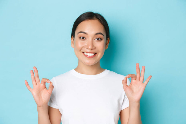 Detailní záběr docela spokojené usměvavé asijské dívky v bílém tričku, ukazující doporučení gesto, ok znamení a kývnutí na souhlas, výběr komplimentu, souhlasím s vámi, stojící světle modré pozadí - Fotografie, Obrázek