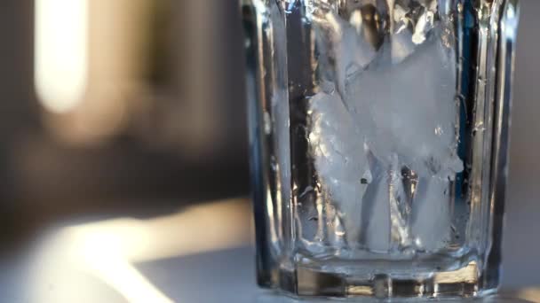 Lanzando trozos de hielo en un vaso transparente, preparando un cóctel. Concepto. Primer plano de cristal de pie sobre la mesa lleno de hielo frío sobre fondo borroso. - Metraje, vídeo