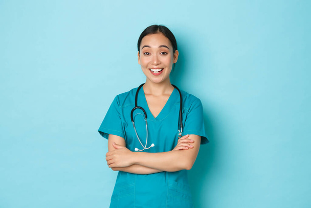 Covid-19, соціальне дистанціювання та концепція пандемії коронавірусу. Весела усміхнена азіатська жінка-лікар, лікар або медсестра в скрабах схрещують руки і обговорюють щось, стоячи на світло-блакитному фоні
 - Фото, зображення