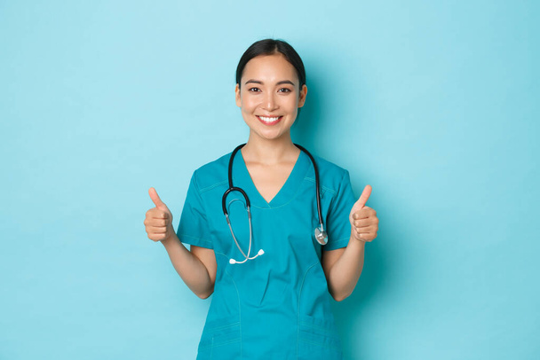 Covid-19, social distancing and coronavirus pandemic concept. Optimistic and supportive asian female doctor, nurse encourage everything okay, thumbs-up in approval and smiling, wearing scrubs - Photo, Image