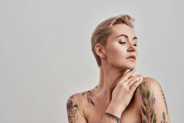 Beauty portrait of a young attractive half naked tattooed woman with perfect skin looking away, holding hand near her face isolated over grey background - Valokuva, kuva