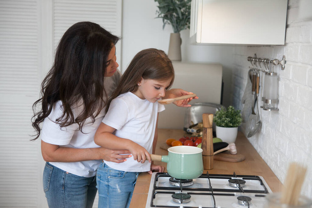 Happy loving family preparing dinner together. Smiling Mom and child daughter girl cooking and having fun in the kitchen. Homemade healthy food. Little helper in the white Scandinavian-style interior - Photo, Image