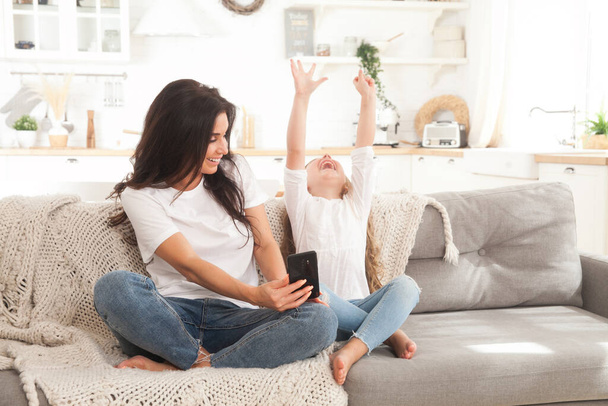 Atrakcyjna młoda mama i córeczka grające w gry online w domu za pomocą smartfona, posiadające wideo czat lub oglądanie filmu. Kształcenie na odległość. Wideokonferencja lub wirtualne wydarzenie związane z kwarantanną - Zdjęcie, obraz