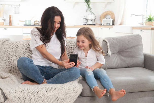 Houkutteleva nuori äiti ja pieni tytär pelaa online-pelejä kotona älypuhelimella, jolla on video chat tai katsomassa elokuvaa. Etäopetus. Videoneuvottelu tai virtuaalinen tapahtuma karanteenissa - Valokuva, kuva