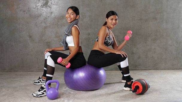 Spor salonunda spor salonunda spor topu ve dambılla poz veren iki güzel Asyalı kadın. Mutlu bayan arkadaşlar boş zamanlarını spor merkezinde geçiriyorlar. - Fotoğraf, Görsel
