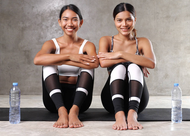 İki güzel Asyalı kadın mola veriyor. Spor salonundaki yoga minderinde su içiyorlar. Mutlu bayan arkadaşlar boş zamanlarını spor merkezinde geçiriyorlar. - Fotoğraf, Görsel