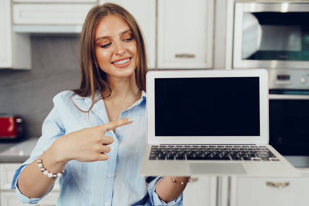 Young blonde woman standing in kithen and showing blank screen laptop computer - Fotoğraf, Görsel