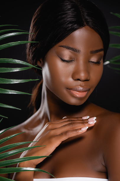 african american naked woman with closed eyes near green palm leaves isolated on black - Foto, imagen