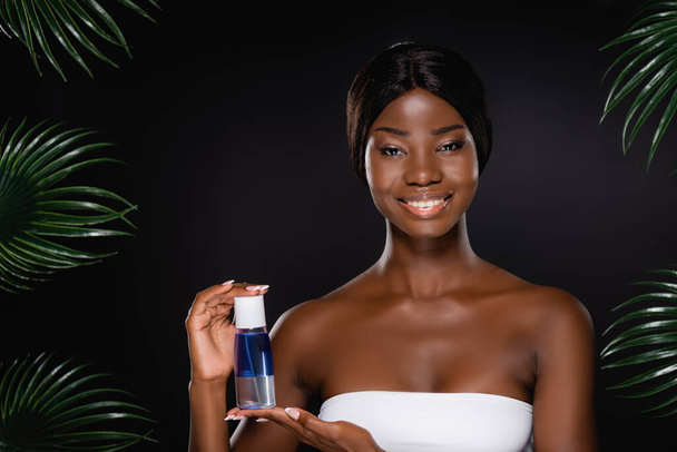 african american woman holding micellar water near green palm leaves isolated on black - Foto, immagini