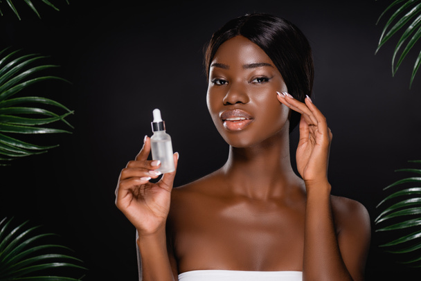 african american woman holding bottle with serum near green palm leaves isolated on black - Photo, Image