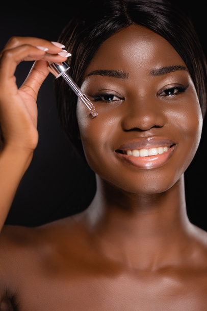portrait of african american naked woman applying serum on face isolated on black - Photo, Image