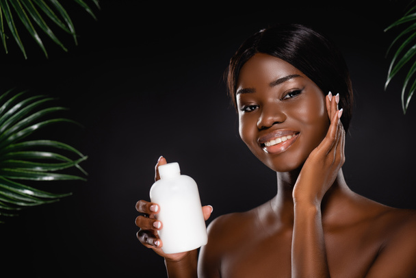 african american naked woman holding bottle of lotion near green palm leaves isolated on black - Photo, Image
