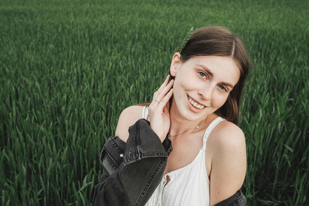 Young Stylish Girl in White Dress and Denim Jacket Spends Time in Green Wheat Field - Photo, image